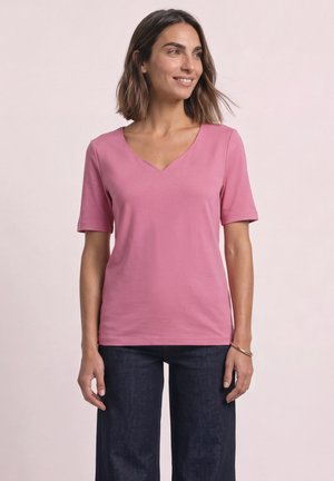 Femme portant un t-shirt rose à col en V manches courtes et un jean foncé, debout devant un fond clair uni, regardant sur le côté et souriant.