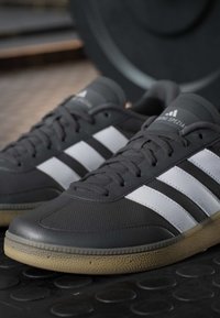 Close-up van zwarte sneakers met witte strepen en rubberen zolen die op een gestructureerde zwarte gymvloer liggen vlakbij een gewichtsschijf.