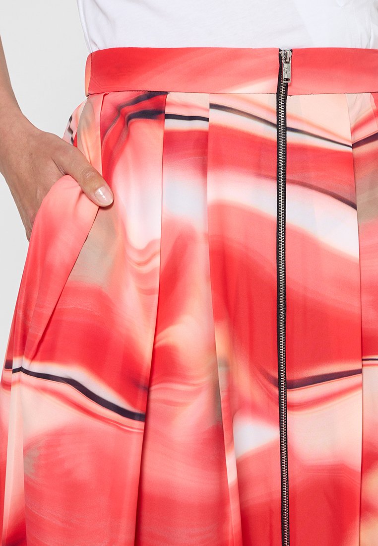 Ręka spoczywa w kieszeni plisowanej spódnicy w abstrakcyjny wzór w kolorach czerwonym, różowym i białym oraz z zamkiem błyskawicznym z przodu.