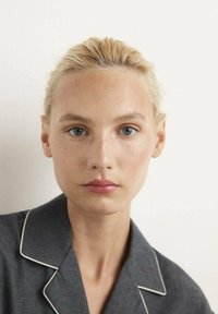 Mujer rubia con ojos azules y pecas claras mira hacia adelante, vistiendo una camisa de color gris oscuro con cuello y ribetes blancos.