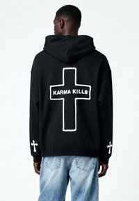 Hoodie nero caratterizzato da un grande ricamo bianco a forma di croce sul retro con la scritta "KARMA KILLS". Accentuato da piccole croci bianche sulle maniche.