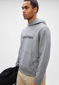 Muž v světle šedé mikině Calvin Klein a béžových kalhotách drží černou bundu na bílém pozadí.