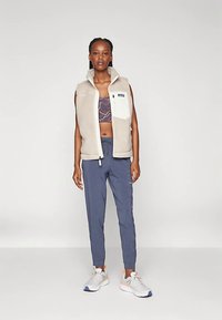 Colete de lã polar em bege claro com fecho frontal e bolso no peito. Usado sobre uma t-shirt curta com padrão e combinado com calças azuis marinho afuniladas e ténis.