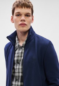 Tumši zils zipa džemperis ar augstu apkakli, izgatavots no gludas auduma; ar melnu un baltu pleda krekla apakšā, piestiprināta logotipa detaļa.