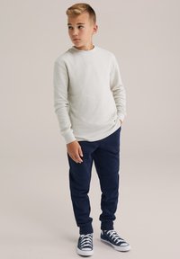 Gaiši pelēks ribots krekls ar garām piedurknēm apvienots ar tumši zilu sporta biksēm un ziliem sporta apaviem. Apģērbs ir brīvā piegriezumā ar elastīgiem rokgriezumiem.