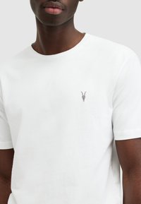 T-shirt bianca in cotone con collo rotondo, maniche corte e un piccolo ricamo di teschio di capra grigio nella zona sinistra del petto.