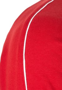 Camiseta atlética roja con una textura suave, que presenta un detalle de ribete blanco a lo largo de la costura del hombro. Vista en primer plano de la tela.