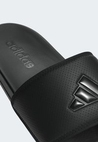 Black Adidas slide sandal featuring a textured strap with a perforated pattern and a raised logo accent. The sole is flat and smooth.