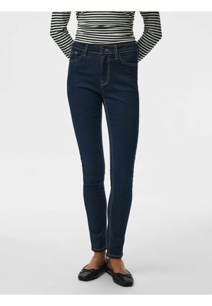 Personne portant un jean skinny bleu foncé, des ballerines noires et un haut à manches longues rayé noir et blanc, les mains dans le dos.