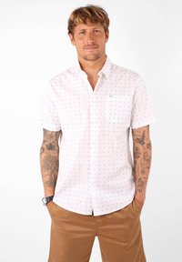 Chemise blanche à manches courtes avec des motifs bleus, design à boutons, poche poitrine et coupe décontractée. Portée avec un short marron clair.