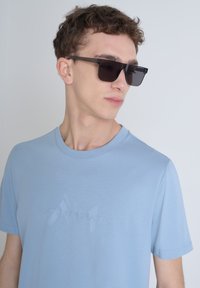 Camiseta de manga corta azul claro con un sutil logo en relieve, combinada con gafas de sol rectangulares negras. Textura suave, diseño casual.
