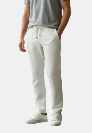 Uomo che indossa una T-shirt grigio chiaro, pantaloni da jogging bianchi a coste con coulisse e scarpe da ginnastica bianche, in piedi con una mano in tasca.