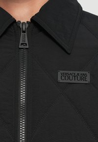 Juodas pūkuotas švarkas su smailiu apykakle, priekiu užtraukiamu užtrauktuku ir matinio juodo logotipo etiketė, pažymėta "VERSACE JEANS COUTURE". Tekstūruoti audinio detalės.