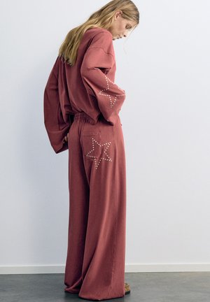 Femme aux longs cheveux portant un pantalon bordeaux ample et un haut à manches longues assorti avec des clous en forme d’étoile sur la manche et la poche arrière.