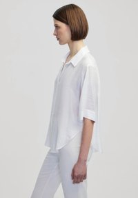 Weiße, leichte Bluse mit kurzen Ärmeln und einem Button-Down-Kragen, mit lässiger Passform und seitlichen Schlitzen, kombiniert mit weißen Hosen.