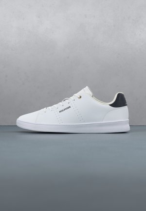 Basket basse blanche avec lacets, languette de talon noire, inscription "HILFIGER" sur le côté et détails perforés sur fond gris.