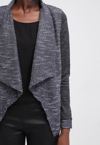 Öppen frontcardigan i grått och svart, med strukturerade stickade mönster, kontrasterande ärmar och draperad design. Mjuk tyg med lös passform.