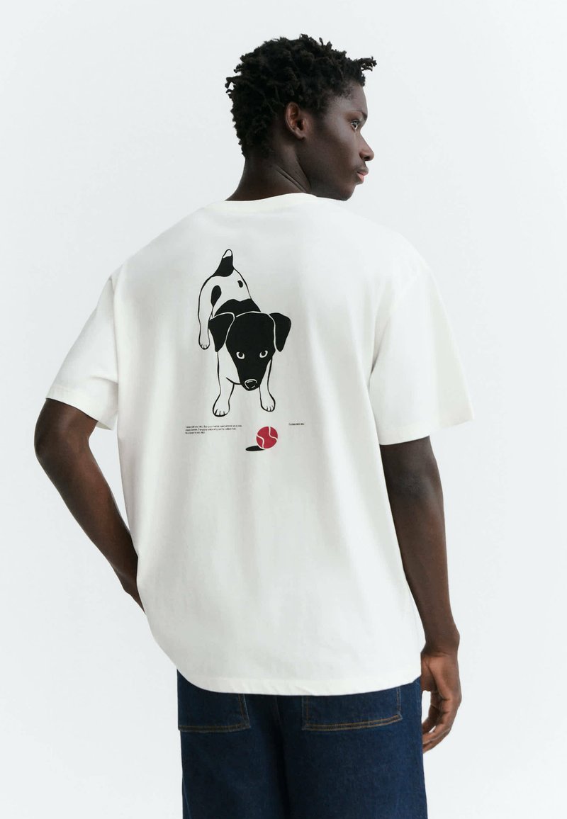 Homme portant un t-shirt blanc avec une illustration en noir et blanc d’un chien poursuivant une balle rouge dans le dos, debout devant un arrière-plan uni.