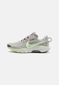 Zapatilla de trail running Nike de color gris claro con un swoosh amarillo pálido, detalles de suela en negro y amarillo, cordones rojos y una pestaña en el talón.