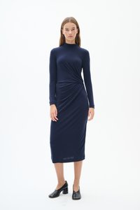 Mörkblå långärmad klänning med en figurnära silhuett, som har en tvinnad detalj vid midjan, tillverkad av mjukt, stretchigt material. Svarta klackskor med blockklack.