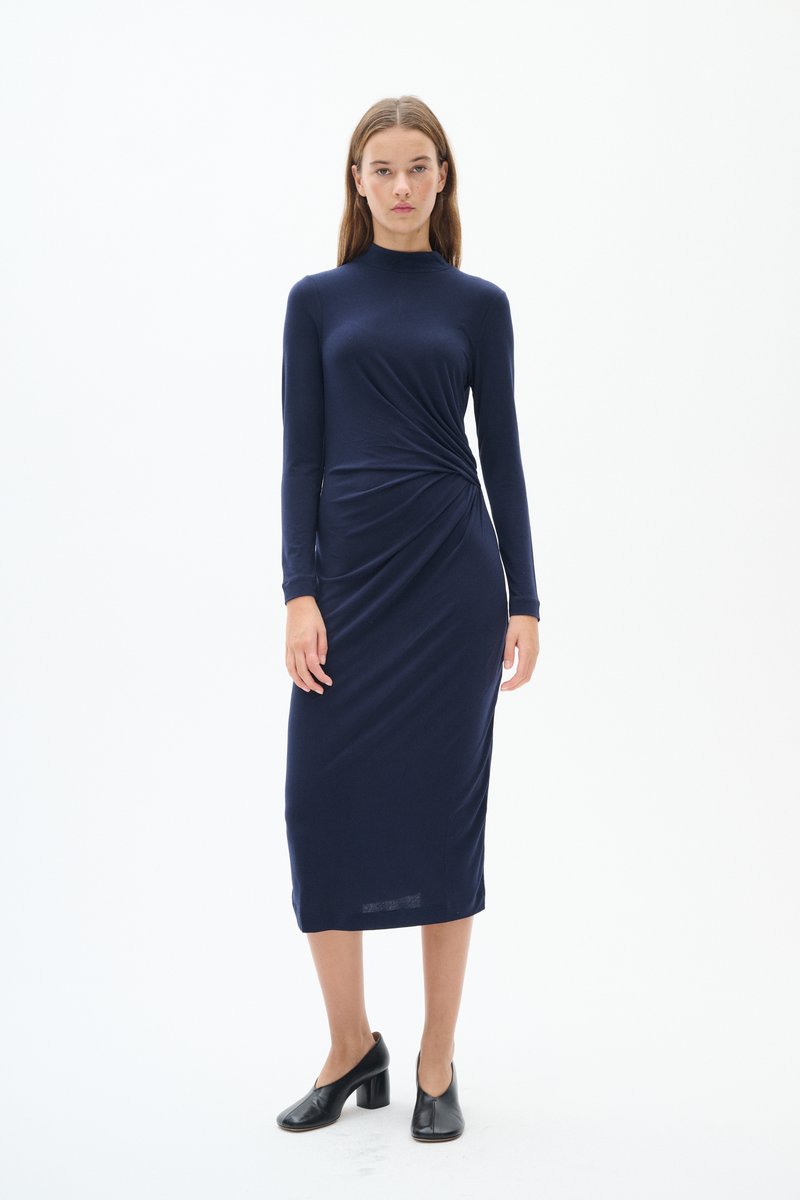 Mörkblå långärmad klänning med en figurnära silhuett, som har en tvinnad detalj vid midjan, tillverkad av mjukt, stretchigt material. Svarta klackskor med blockklack.