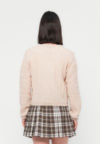 Femme avec des cheveux noirs raides portant un pull en tricot rose pâle et une jupe à carreaux plissée, se tenant face à un fond blanc.