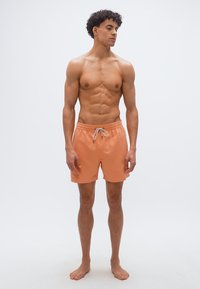 Μυώδης νεαρός με σγουρά μαλλιά που φοράει πορτοκαλί μαγιό, στέκεται ξυπόλυτος μπροστά σε απλό λευκό φόντο, κοιτώντας προς το πλάι.