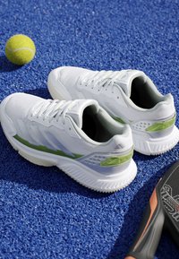 Scarpe sportive bianche e verdi su un campo da tennis blu vicino a una pallina da tennis gialla e parte di una racchetta nera da paddle.