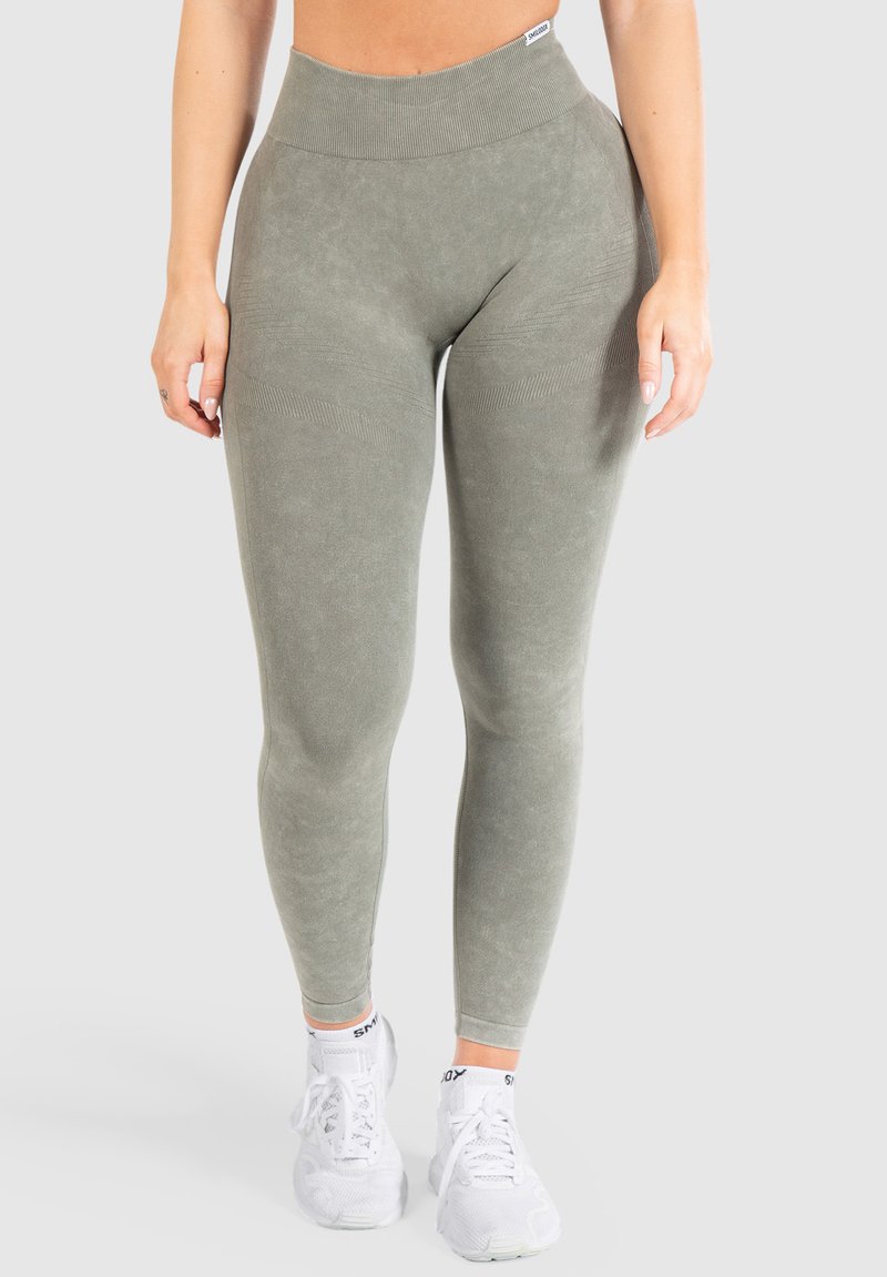Leggings a vita alta, aderenti, di un verde tenue, in tessuto testurizzato con cintura a coste e dettagli sottili sulle gambe. Sneakers bianche visibili.