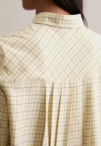 Light yellow collared shirt with a grid pattern of thin, dark lines. Features pleats and a soft texture.