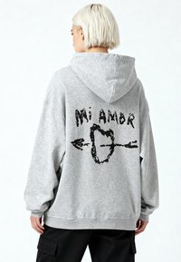 Pernas šviesiai pilka džemperis su didelėmis rankovėmis, pagamintas iš medvilnės mišinio, turi juodą grafikos spaudą su užrašu „Mi Amor“, širdimi ir strėlėmis ant nugaros.