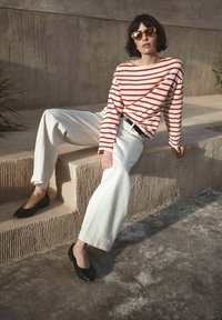 Frau mit kurzen dunklen Haaren, die herzförmige Sonnenbrillen, ein rot-weiß gestreiftes Oberteil, weiße Hosen und schwarze Ballerinas trägt und auf Betontreppen sitzt.