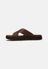 Braune Lederschlappe mit gekreuzten Riemen und gepolsterter Sohle, seitlich auf einem einfarbigen Hintergrund gezeigt.