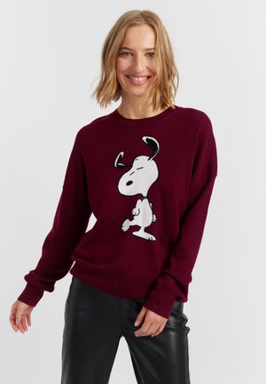 Bordeaux brei sweater met een witte Snoopy-afbeelding in een speelse pose. Losse pasvorm met geribde boorden en zoom. Materiaal lijkt zacht te zijn.