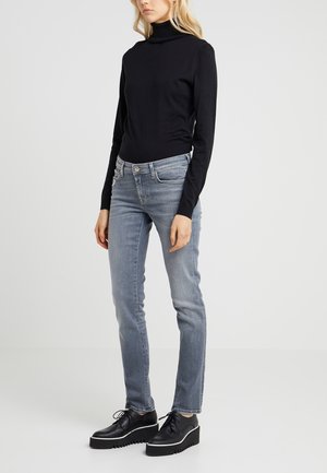 Kvinde iført sort rullekrave, lyseblå slim-fit jeans og sorte platform snøresko står mod en ensfarvet baggrund.