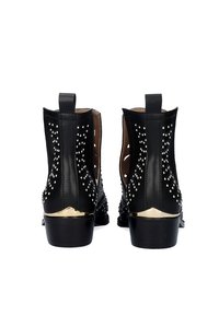 Botas de tobillo de cuero negro con adornos de remaches, laterales con recortes y un tacón con acentos dorados, que muestra una superficie texturizada.
