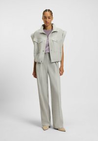 Vaaleanharmaa ylisuuri liivi, jossa on korkea kaulus ja napit, yhdistettynä samanvärisiin leveisiin housuihin. Liivissä on kaksi etutaskua ja sileä rakenne.