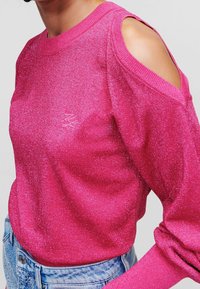 Frau in einem glitzernden pinken Cold-Shoulder-Pullover, kombiniert mit hellblauen Jeans aus Denim mit Verzierungen.