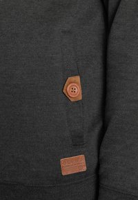 Mørkegrå sweatshirt i et teksturert stoff, med en fremtredende lærpatch og knappdetalj på en sidelomme. Merkelapp synlig.