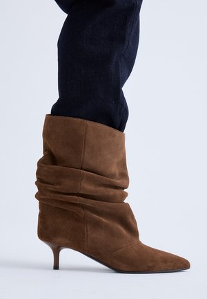 Botas marrones de ante hasta media pantorrilla con punta afilada y tacones pequeños, usadas con pantalones oscuros sobre un fondo liso.