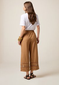 Pantalon large marron avec une taille élastique et un ourlet détaillé. Porté avec un t-shirt blanc et des sandales noires. Texture lisse et coupe décontractée.