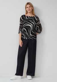 Schwarze und beige transparente Bluse mit gewellten Mustern, Puffärmeln, kombiniert mit lockeren schwarzen Hosen und weißen Sneakers. Schlichte Accessoires.