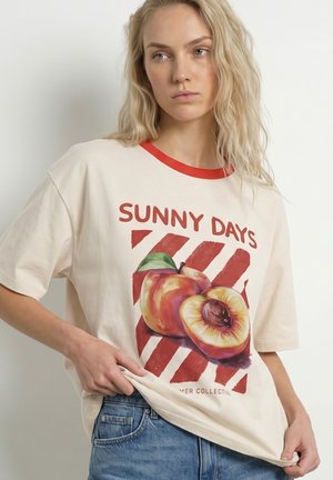 Jeune femme aux longs cheveux blonds porte un t-shirt crème avec un liseré rouge et un motif pêche intitulé "Sunny Days" ainsi qu'un jean bleu.
