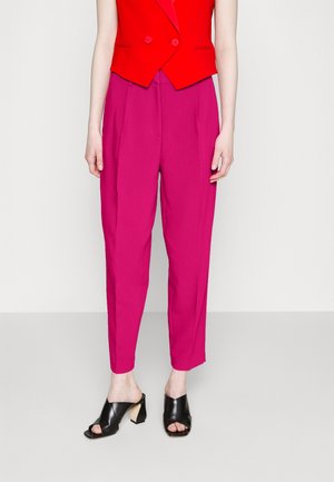 Femme portant un pantalon rose vif fuselé, un gilet rouge court croisé et des mules à talons noires ouvertes au bout.