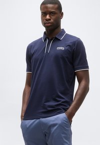 Άνδρας που φοράει μπλε ναυτικό πόλο πουκάμισο Calvin Klein με λευκή διακόσμηση και ανοιχτό μπλε παντελόνι, στέκεται με τα χέρια στις τσέπες.