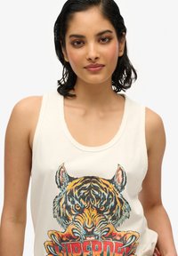 Débardeur crème avec un graphique détaillé et coloré d'un tigre et le mot "Superdry" en lettres sobres, avec une coupe décontractée et une texture douce.