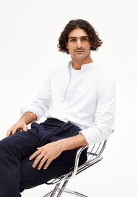 Weißes Hemd mit Stehkragen, kombiniert mit marineblauen Hosen. Die Figur sitzt auf einem chromfarbenen Stuhl, die Hände auf den Oberschenkeln ruhend.