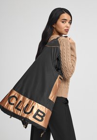 Svart och bronzfärgad vindjacka med stor text "CLUB". Har en glänsande bronzefärgad detalj och en lätt design, med kviltad textur.