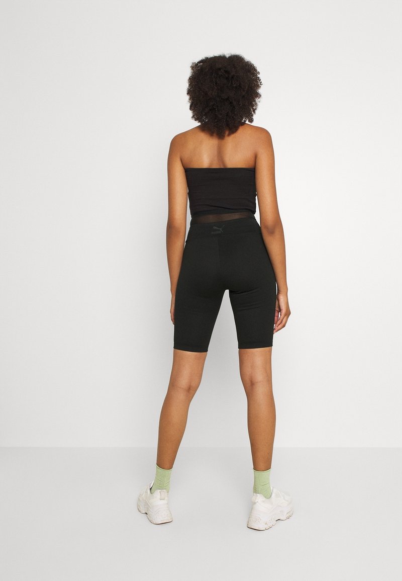 Femme aux cheveux bouclés vue de dos, portant un haut bandeau noir, un short de cyclisme ajusté noir, des chaussettes vert clair et des baskets blanches.