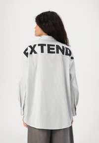 Camicia a righe a maniche lunghe di colore grigio chiaro con orlo curvo. Presenta un audace testo nero "EXTEND." sulla parte posteriore e polsini con bottoni.